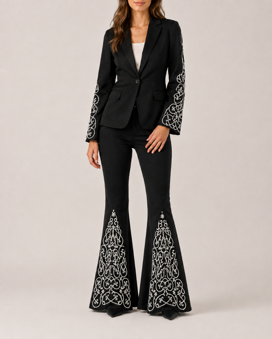 A model standing against a solid backdrop is seen wearing black embroidered flare jeans with a matching black embroidered blazer with embroidered sleeves.