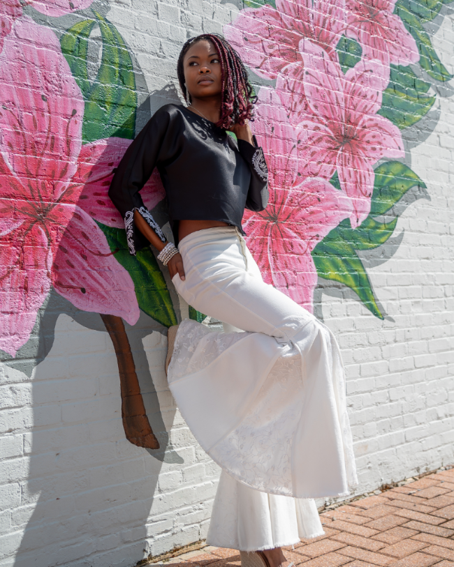 A woman posing against a white brick wall with a large pink floral mural, wearing a black long-sleeve crop top and white wide-leg pants.