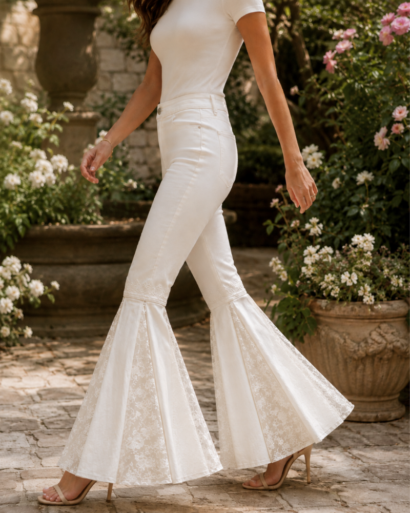 A woman wearing white high-waisted flared pants with lace detailing at the cuffs, paired with a white fitted top and nude heels, standing in a garden courtyard with stone paving and flower planters.