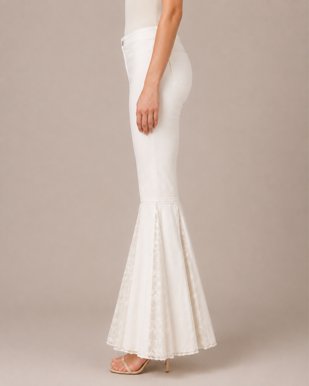 A model shown from the waist down wears a white high-waisted mermaid-style skirt with a sheer lace flared hem, paired with nude high heels, against a solid beige studio background.