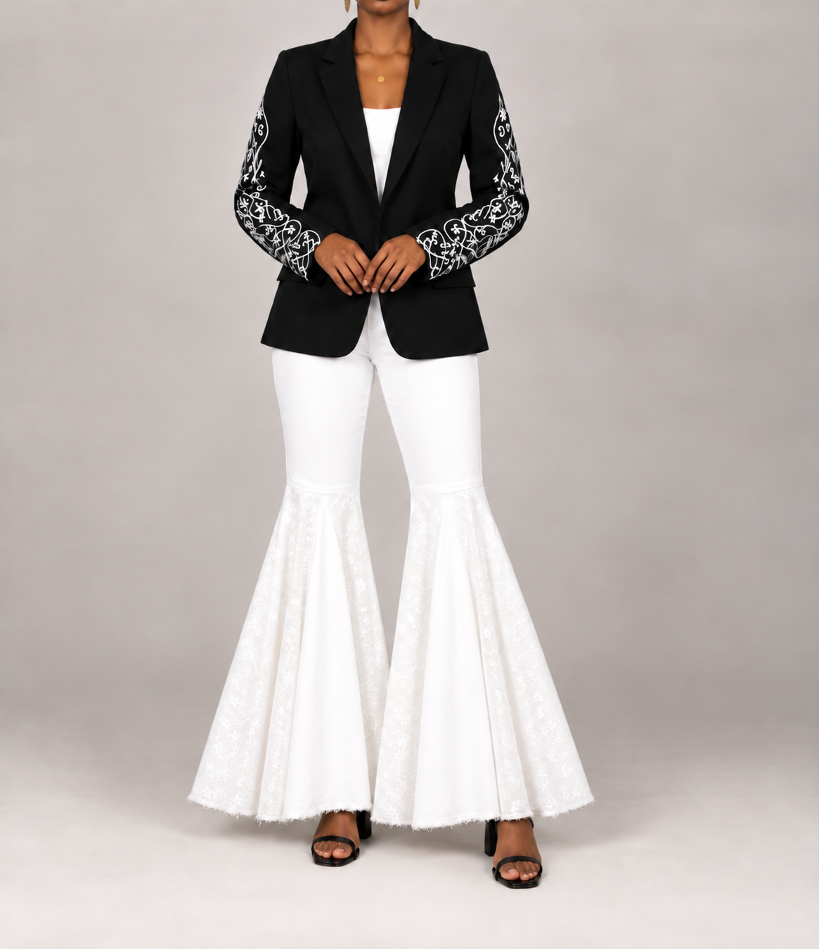 A model wearing a black blazer with intricate white floral embroidery on the sleeves, paired with white flared pants and black sandals, against a neutral gray studio background.