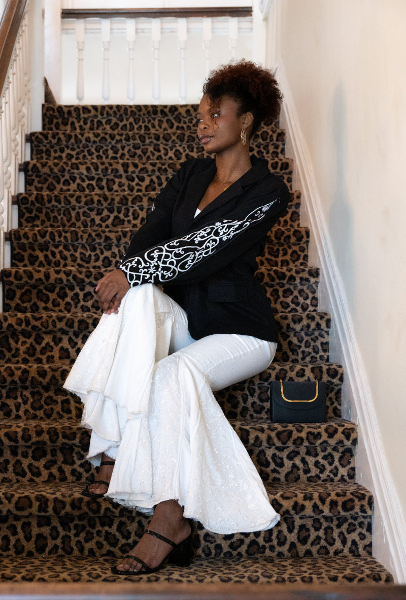 A woman sits on a staircase wearing a black blazer with white embroidered sleeve detailing, white flared pants, and black sandals. A small black handbag with gold trim rests beside her on the steps.